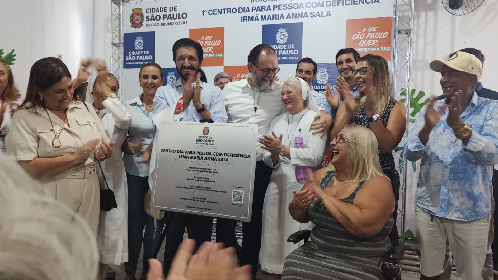 Na imagem pessoas estão reunidas ao redor da placa de inauguração do Centro Dia para Pessoas com Deficiência Irmã Maria Anna Sala. Ao centro, atrás da placa, temos o Prefeito de São Paulo, Ricardo Nunes, e o Secretário de Assistência e Desenvolvimento Social, Carlos Bezerra Jr. Todos aplaudem e sorriem, comemorando a inauguração do espaço.